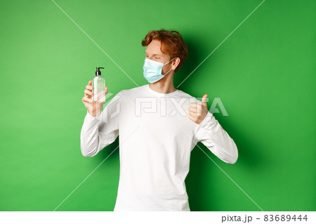 Covid-19, virus and social distancing concept. Handsome young man with red hair, wearing medical mask, looking at antiseptic and showing thumb-up, green background 83689444