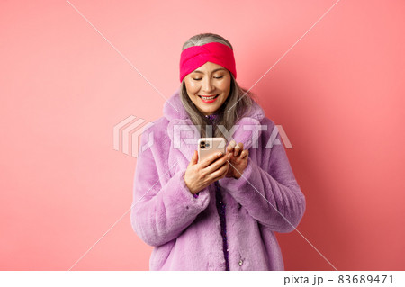 Online shopping and fashion concept. Modern asian granny in trendy faux fur coat texting message, using smartphone and looking happy at screen, pink background Online shopping and fashion concept. Modern asian granny in trendy faux fur coat texting message, using smartphone and looking happy at screen, pink background 83689471