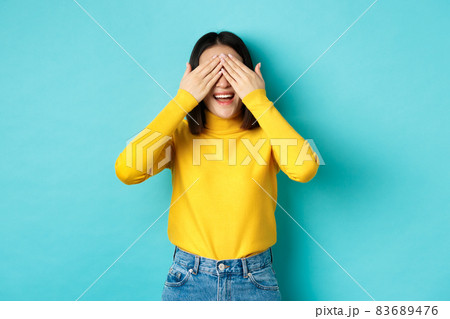 Cheerful asian girl in yellow pullover waiting for surprise, playing hide n seek and smiling, expecting gift with eyes closed, standing over blue background Cheerful asian girl in yellow pullover waiting for surprise, playing hide n seek and smiling, expecting gift with eyes closed, standing over blue background 83689476