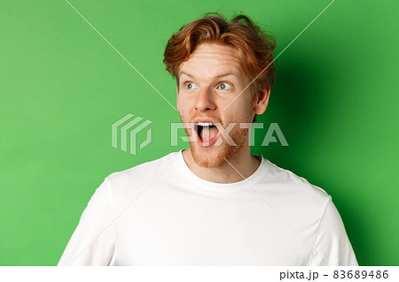Close up of surprised and impressed redhead man checking out promotion offer, looking left with dropped jaw, standing over green background Close up of surprised and impressed redhead man checking out promotion offer, looking left with dropped jaw, standing over green background 83689486