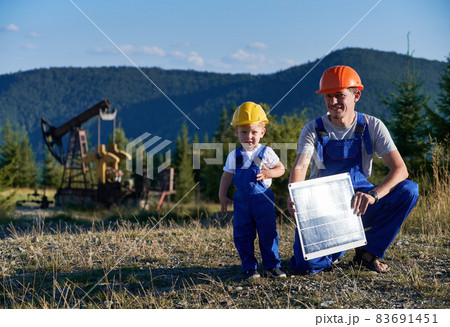 Portrait of professional Caucasian mechanic with solar panel learning capable boy derrick maintenance, father and son dressed in blue uniform looking at camera during daytime at petroleum station 83691451