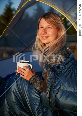 Portrait of female hiker resting in tourist tent with happy expression, enjoying tasty morning coffee. Camping in nature. Active hike with overnight stay in tent camp. Portrait of female hiker resting in tourist tent with happy expression, enjoying tasty morning coffee. Camping in nature. Active hike with overnight stay in tent camp. 83699256