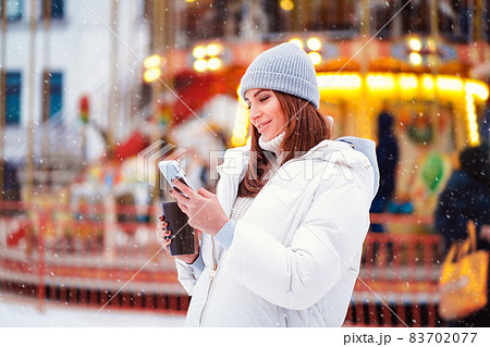 Side view of happy woman wears in jackets reads smartphone at the Christmas city while holds coffe Side view of happy woman wears in jackets reads smartphone at the Christmas city while holds coffe 83702077