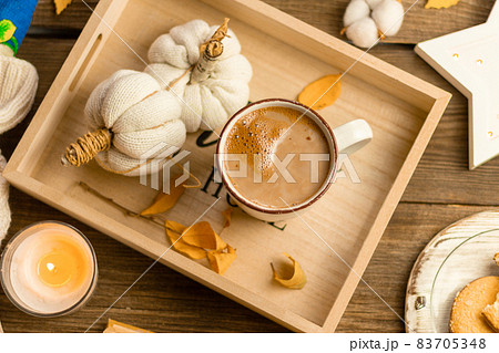 Autumn still life of a wooden tray, white knitted handmade pumpkins, yellow leaves, a mug of cocoa, coffee or hot chocolate, lit candles, round cookies. Warm home comfort concept 83705348