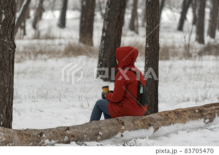 Woman in red warm clothes sits on log in winter park with cup of coffee in her hands. Walk through the winter forest. 83705470