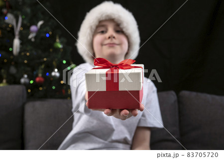 Boy in Santa hat is holding out present to you sitting on the sofa near the Christmas tree. New Year. Christmas. Boy in Santa hat is holding out present to you sitting on the sofa near the Christmas tree. New Year. Christmas. 83705720