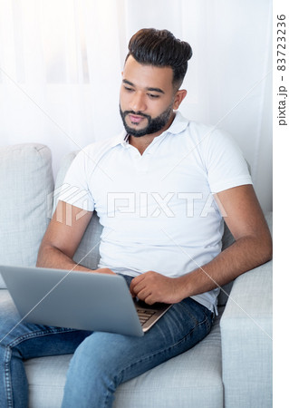 Online communication. Happy man. Home relax. Handsome arabian guy sitting comfortable sofa with laptop in light room interior. Online communication. Happy man. Home relax. Handsome arabian guy sitting comfortable sofa with laptop in light room interior. 83723236