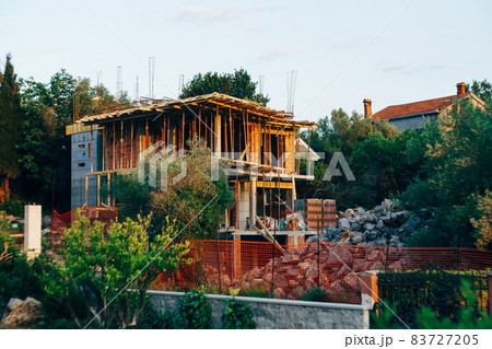 Multi-storey house under construction behind a fence against a background of green trees 83727205