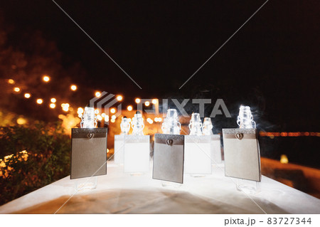 Sealed bottles with blank signs stand on a table covered with a white tablecloth against a backdrop of glowing light bulbs at night Sealed bottles with blank signs stand on a table covered with a white tablecloth against a backdrop of glowing light bulbs at night 83727344