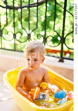 Little baby with a dipper in his hands sits in a basin with water and toys 83727507