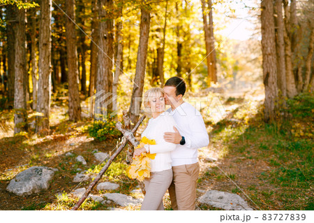 Man kisses and hugs smiling woman by the shoulders. The woman has yellow leaves in her hands. Wide angle 83727839