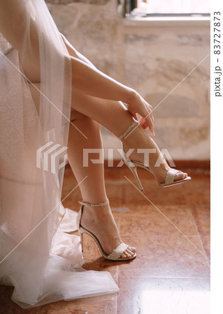 Bride in a white dress buttons up her high-heeled sandals. Close-up 83727873