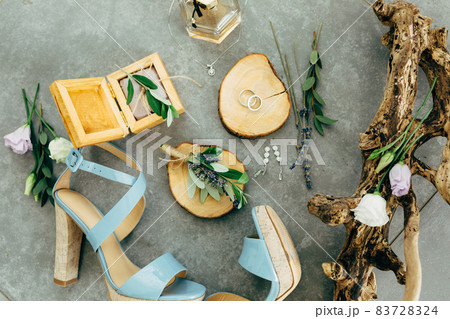 Wedding rings lie in a wooden stand surrounded by sandals with heels, wooden box, perfume, flowers and twigs on the floor 83728324