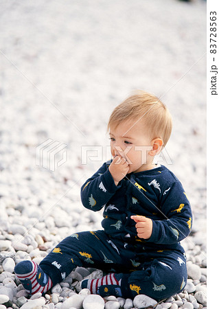 Cute baby in blue overalls sits on a pebble beach and holds a pebble in his hand, which he puts in his mouth Cute baby in blue overalls sits on a pebble beach and holds a pebble in his hand, which he puts in his mouth 83728563