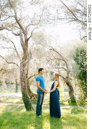 Tall man in a blue T-shirt holds the hands of pregnant woman in a long dress against the backdrop of huge green olive trees Tall man in a blue T-shirt holds the hands of pregnant woman in a long dress against the backdrop of huge green olive trees 83729002