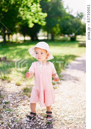 Little girl in a hat walks along the gravel path in the park with a stick in her hand Little girl in a hat walks along the gravel path in the park with a stick in her hand 83730001