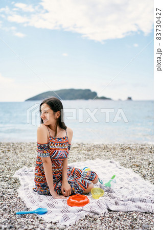 Smiling girl sitting on a blanket on a pebble beach, turning her head to the side 83730427