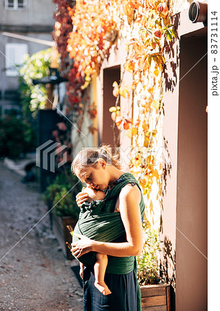 Mom hugs the baby in a sling near the wall of the house, entwined with ivy 83731114
