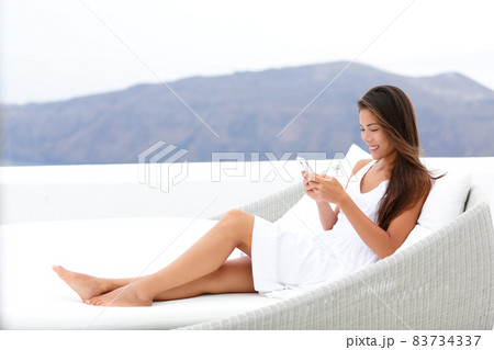 Luxury lifestyle woman relaxing in outdoor sofa using her mobile phone texting sms text message online sitting in chair lounger on hotel balcony - home comfort. 83734337
