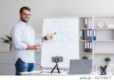 Smiling adult european man teacher in glasses shows english rules on board and looks at smartphone webcam Smiling adult european man teacher in glasses shows english rules on board and looks at smartphone webcam 83734877