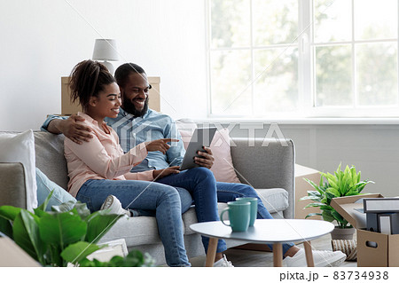 Satisfied young african american man and woman sit on sofa, rest among boxes and plants after moving 83734938