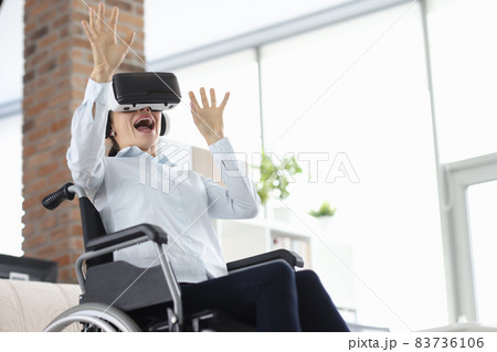 Woman sitting in wheelchair wearing virtual reality glasses and waving her hands 83736106
