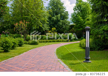 lantern iron ground garden lighting of a park path paved with stone tiles in the backyard among plants, bushes and trees surrounded by a green lawn on a sunny summer day, nobody. 83736225