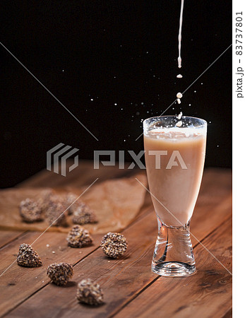 A stream of milk is poured into a glass with a milk drink with a splash of drops. Chocolate candies lie on a wooden table A stream of milk is poured into a glass with a milk drink with a splash of drops. Chocolate candies lie on a wooden table 83737801