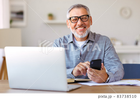 Smiling senior man using mobile phone while sitting at his cozy workplace with laptop at home Smiling senior man using mobile phone while sitting at his cozy workplace with laptop at home 83738510