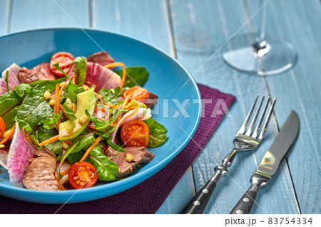 Spicy beef slices meat salad with tomatoes, parsley, radish and salad leaves spinach in a blue plate on a wooden table. Spicy beef slices meat salad with tomatoes, parsley, radish and salad leaves spinach in a blue plate on a wooden table. 83754334