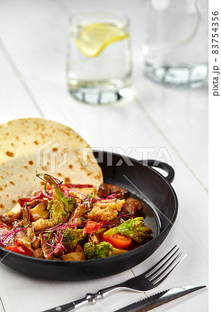 Roasted vegetables - potato, green beans, carrot and paprika in black iron plate on white wooden table. 83754356