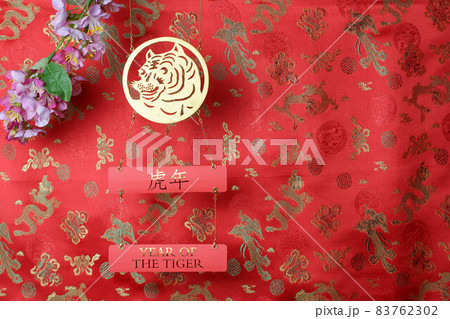 Golden tiger symbol and branch of pink sakura cherry blossom on red fabric with traditional chinese ornament birds and dragons. Chinese New Year of the Tiger 2022. Translation: year of the tiger Golden tiger symbol and branch of pink sakura cherry blossom on red fabric with traditional chinese ornament birds and dragons. Chinese New Year of the Tiger 2022. Translation: year of the tiger 83762302