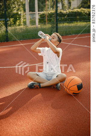 Multiracial boy sitting on a basketball court with a bottle of water Multiracial boy sitting on a basketball court with a bottle of water 83762376
