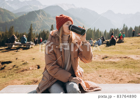 Young blond woman with thermos cup resting on the bench in mountains. Freedom, food, travel and vacations concept, outdoor activities. Happy Girl wearing red hat, halt 83764890