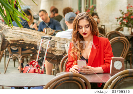 Tourist young caucasian white woman in a red jacket looking at smartphone sitting at the table in cafe outdoors. Tourist young caucasian white woman in a red jacket looking at smartphone sitting at the table in cafe outdoors. 83766526
