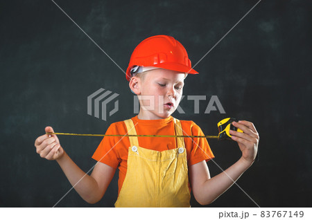 Portrait of child in safaty red helmet and builder uniform as a construction worker with measuring tape. School kid boy dreams of becoming builder guy Children profession. Smart kid 83767149