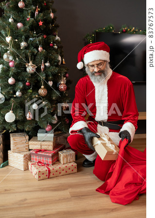 Smiling bad Santa in black gloves crouching at decorated tree and stealing Christmas presents Smiling bad Santa in black gloves crouching at decorated tree and stealing Christmas presents 83767573