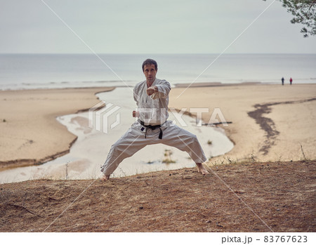 Karate man in an old kimono and black belt training punch in kibadachi stand at the sea. Martial arts concept. The river flows into the sea at the background.  83767623