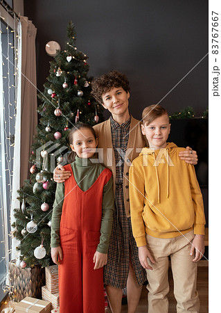 Portrait of smiling middle-aged Caucasian woman in knitted vest embracing kids against Christmas tree at home 83767667