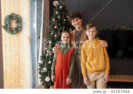 Portrait of smiling middle-aged woman with short curly hair embracing two children against Christmas tree Portrait of smiling middle-aged woman with short curly hair embracing two children against Christmas tree 83767668