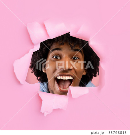 Excited African American teen opening mouth in shock, shouting WOW, looking through hole in ripped pink paper 83768813