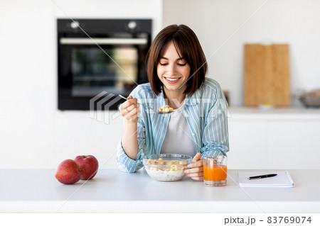 Young lady eating healthy oatmeal, sitting at modern kitchen interior, enjoying granola with milk for breakfast 83769074