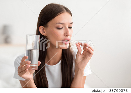 Millennial pretty caucasian female hold pill and glass of water in bedroom interior Millennial pretty caucasian female hold pill and glass of water in bedroom interior 83769078