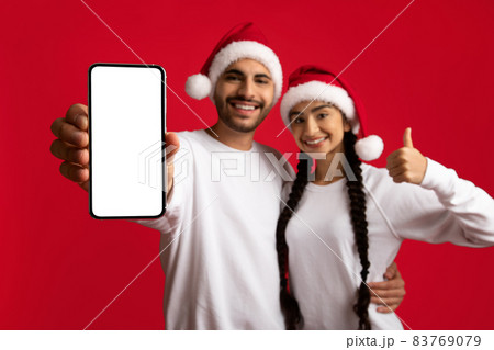 Christmas Offer. Happy Arab Couple Wearing Santa Hats Demonstrating Blank Smartphone Screen Christmas Offer. Happy Arab Couple Wearing Santa Hats Demonstrating Blank Smartphone Screen 83769079