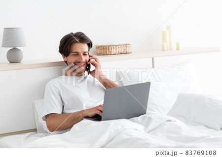 Cheerful attractive millennial european man works on laptop, speaks on phone with client on bed Cheerful attractive millennial european man works on laptop, speaks on phone with client on bed 83769108