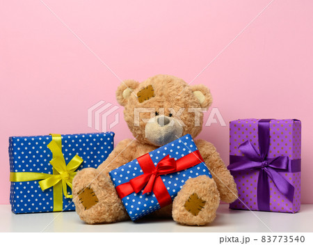 cute brown teddy bear holding a box wrapped in paper and red silk ribbon on white table. Prize and congratulations, pink background cute brown teddy bear holding a box wrapped in paper and red silk ribbon on white table. Prize and congratulations, pink background 83773540