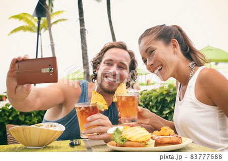 Selfie photo couple taking picture with phone at Hawaii beach bar on summer vacation. Happy Asian woman and man toasting mai tai drinks at bar having fun. Friends on travel holidays. 83777688
