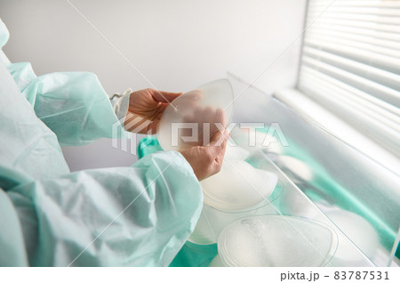 Breast prothesis in surgeon hands on background of medical samples of breast silicone implants of various sizes for breast augmentation aesthetic plastic surgery after resection due to cancer disease 83787531