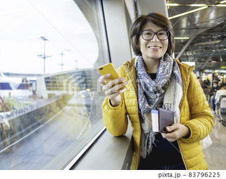 Smiling woman with passport and boarding pass is texting on her smartphone. Tourist is waiting for boarding at window at airport. 83796225