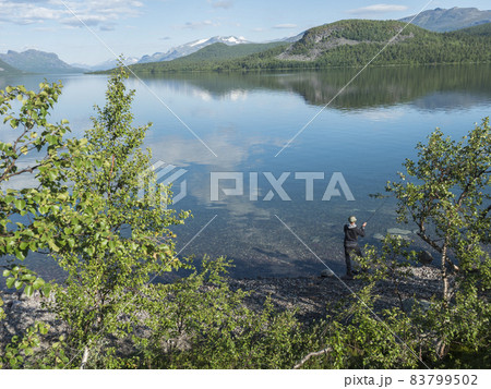 Fisherman at Lule river, in clear calm Lulealven dam lake fresh water in Saltoluokta in Sweden Lapland at sunny summer day. Green rocky mountain, birch trees and clouds. Fisherman at Lule river, in clear calm Lulealven dam lake fresh water in Saltoluokta in Sweden Lapland at sunny summer day. Green rocky mountain, birch trees and clouds. 83799502